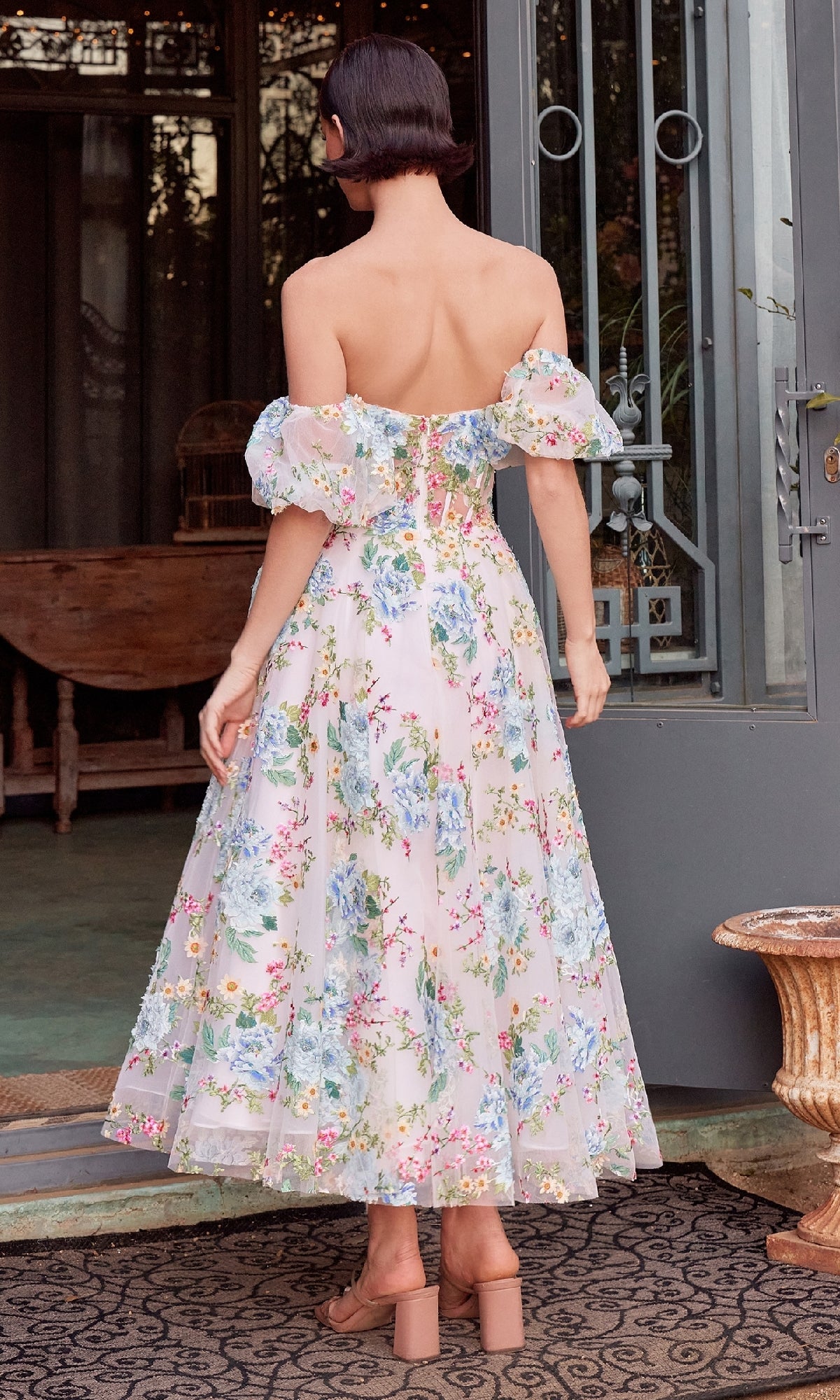 A woman stands with her back to the camera wearing the Andrea & Leo A1406 Floral Prom Dress, featuring removable puff sleeves and a strapless corset bodice, paired with nude block-heeled sandals near a doorway.